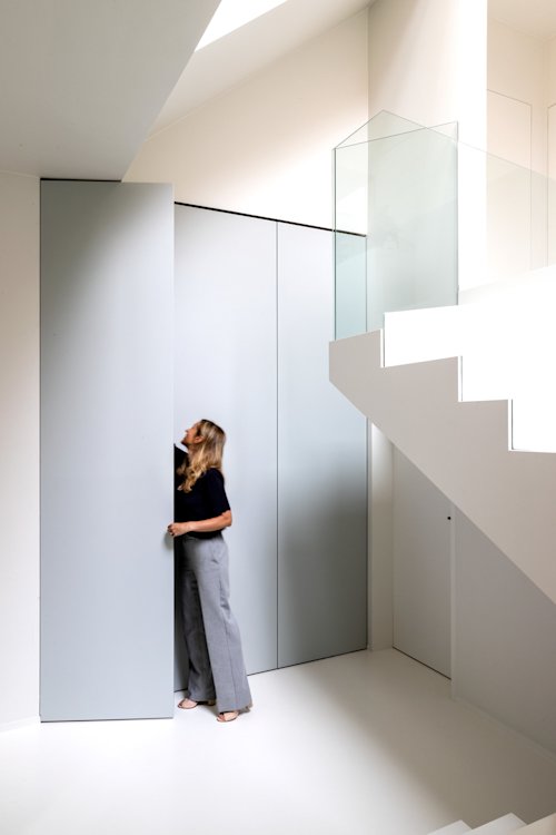 Een vrouw staat in een minimalistisch, wit interieur en reikt omhoog om een hoge, lichtgrijze verborgen deur te openen naast een moderne trap met glazen leuning.