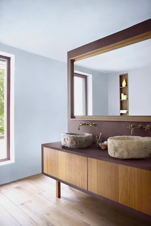 Salle de bains moderne avec meuble en bois, deux grands lavabos en pierre, robinets muraux en cuivre, grand miroir et étagères avec objets décoratifs près de la fenêtre.