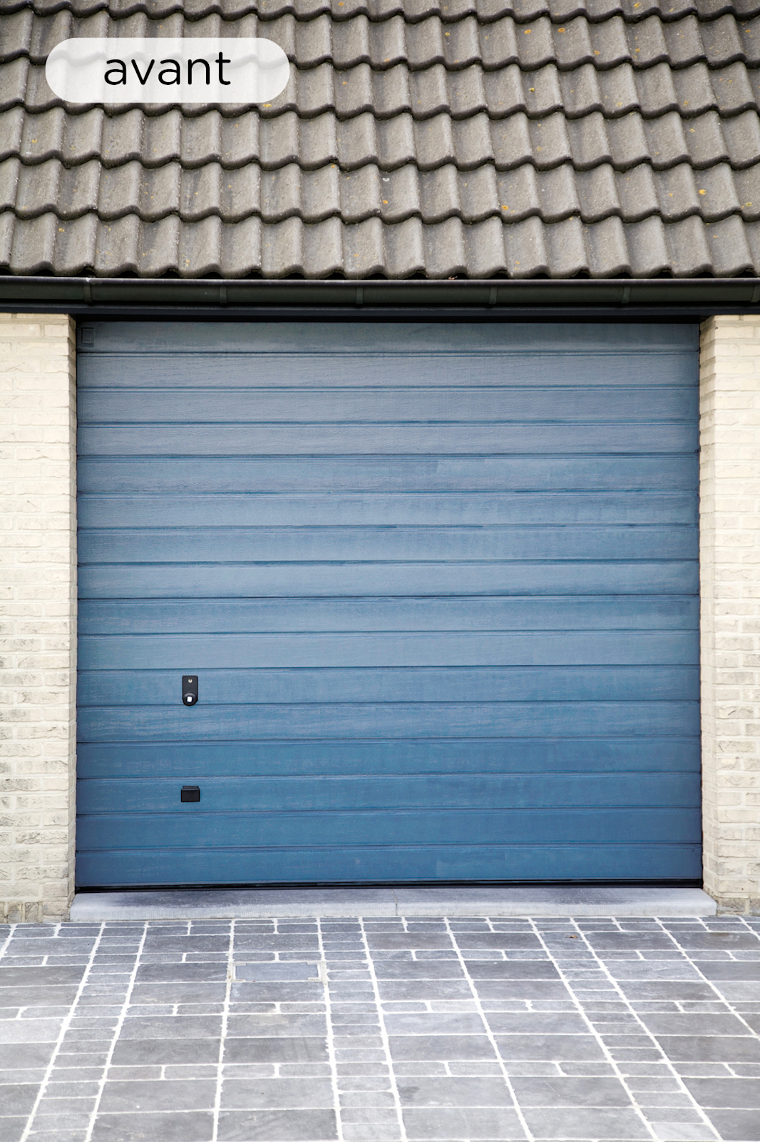 Porte de garage bleue fermée avec des panneaux horizontaux, encadrée de murs en briques claires, sous un toit en tuiles grises et devant une allée pavée en pierre.