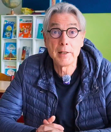 Een oudere man met grijs haar en een bril, gekleed in een blauw jasje, zit aan een tafel. Op de achtergrond staan boekenkasten met kleurrijke boeken. Hij kijkt recht in de camera met zijn handen gevouwen.