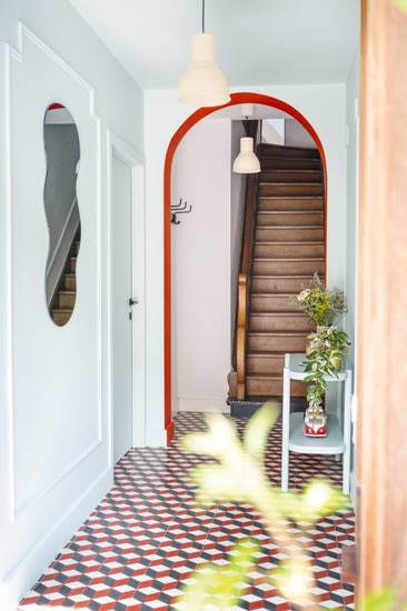 Een hal met vloertegels met geometrische patronen, een rode gewelfde deuropening, een houten trap, een witte wandspiegel en een tafeltje met een vaas bloemen.