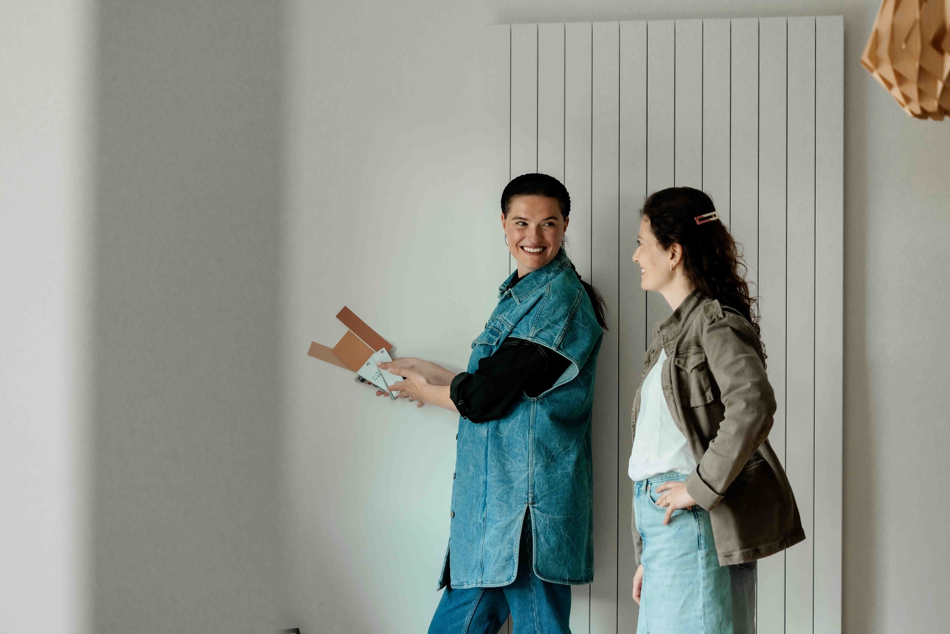 Twee vrouwen staan binnen, de ene houdt kleurstalen vast en lacht naar de andere, die haar hand op haar heup heeft.