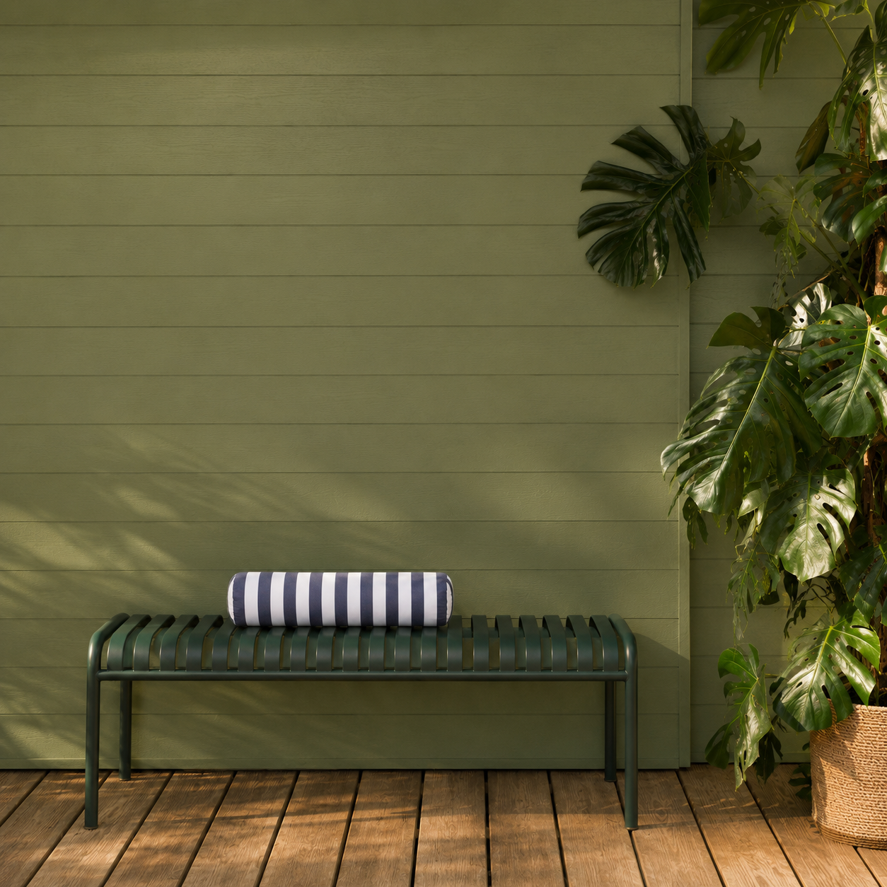 Op een houten terras, tegen een groene muur en naast grote, weelderige planten, staat een groene metalen bank met daarop een opgerolde, gestreepte handdoek.
