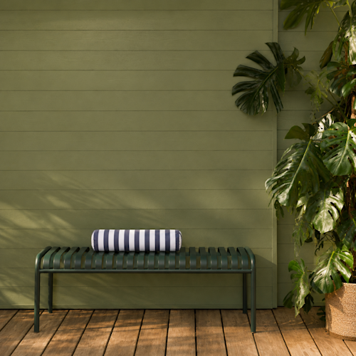 Sur une terrasse en bois, adossé à un mur végétal et à côté de grandes plantes luxuriantes, se trouve un banc en métal vert sur lequel est posée une serviette rayée enroulée.