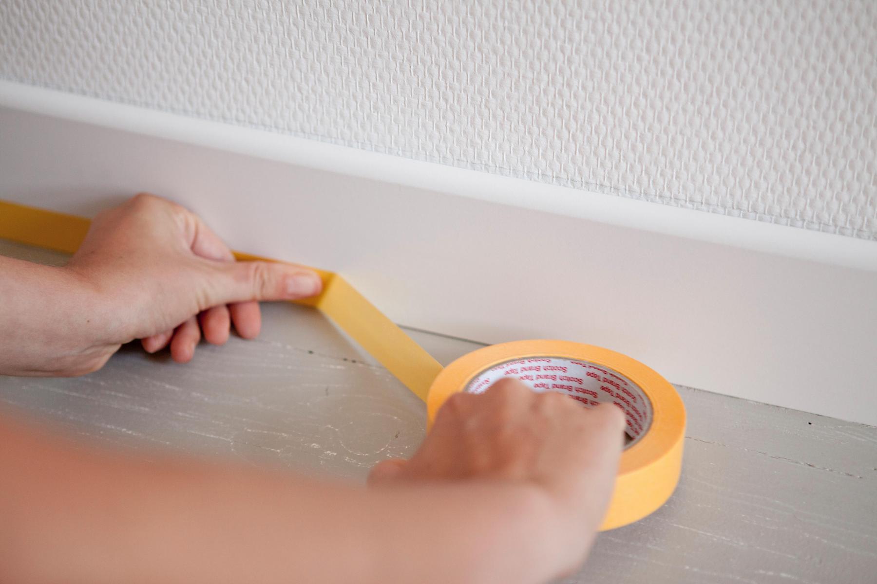 Iemand gebruikt geel afplakband om de rand tussen een witte muurplint en een houten vloer zorgvuldig af te plakken, zodat het gebied klaar is om geschilderd te worden.