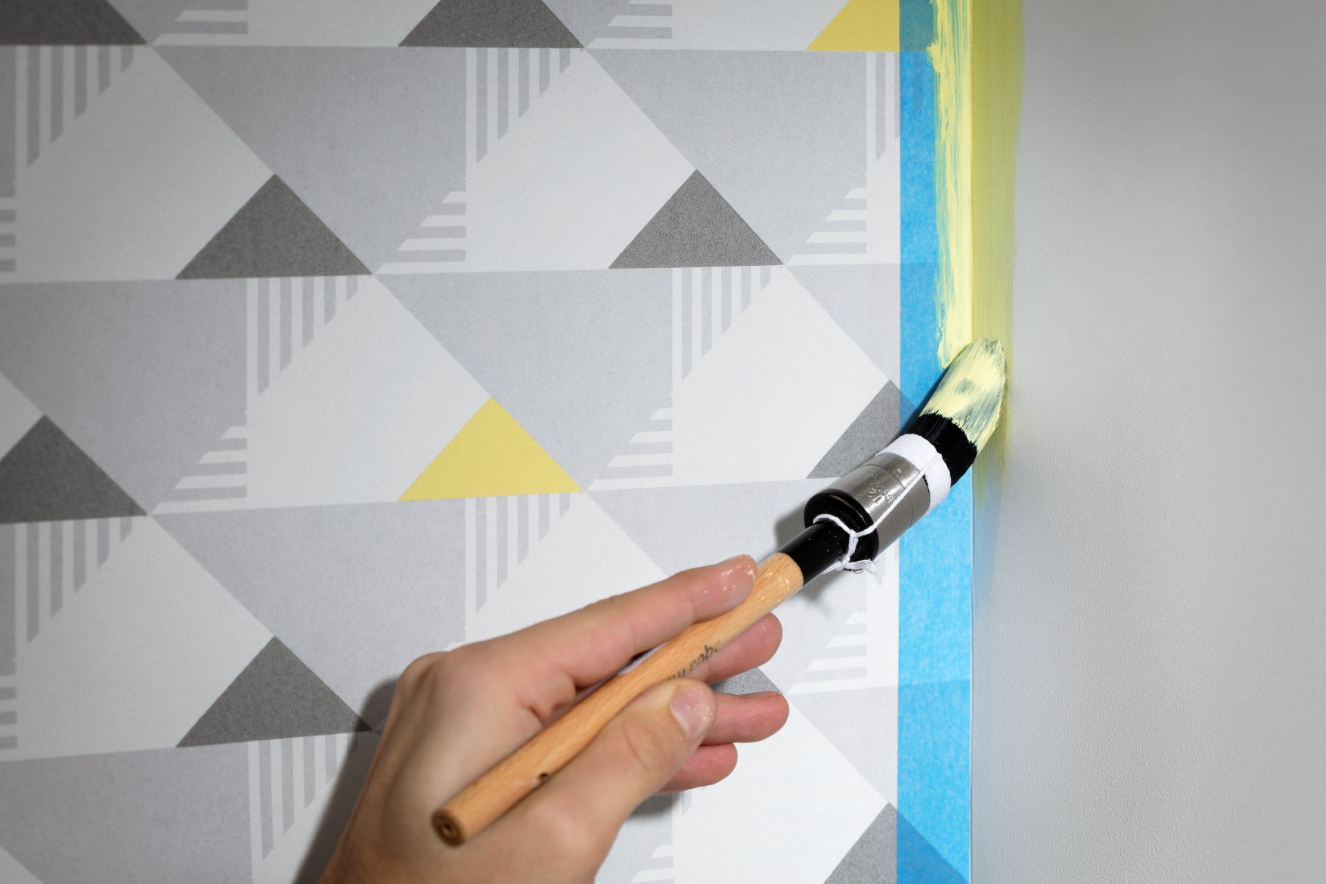 Une main utilise un pinceau pour appliquer de la peinture jaune sur le bord d'un mur recouvert de papier peint à motifs géométriques, en utilisant du ruban adhésif de peintre pour protéger le bord.