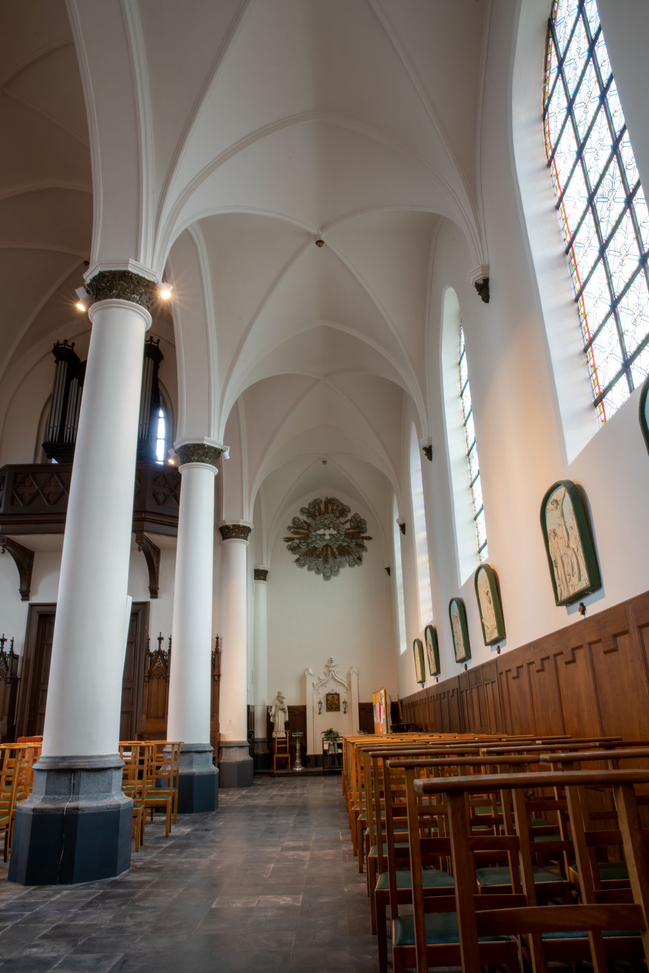 Interieur van een kerk met gewelfde plafonds, hoge witte zuilen, houten kerkbanken, glas-in-loodramen en religieuze kunst aan de muren.