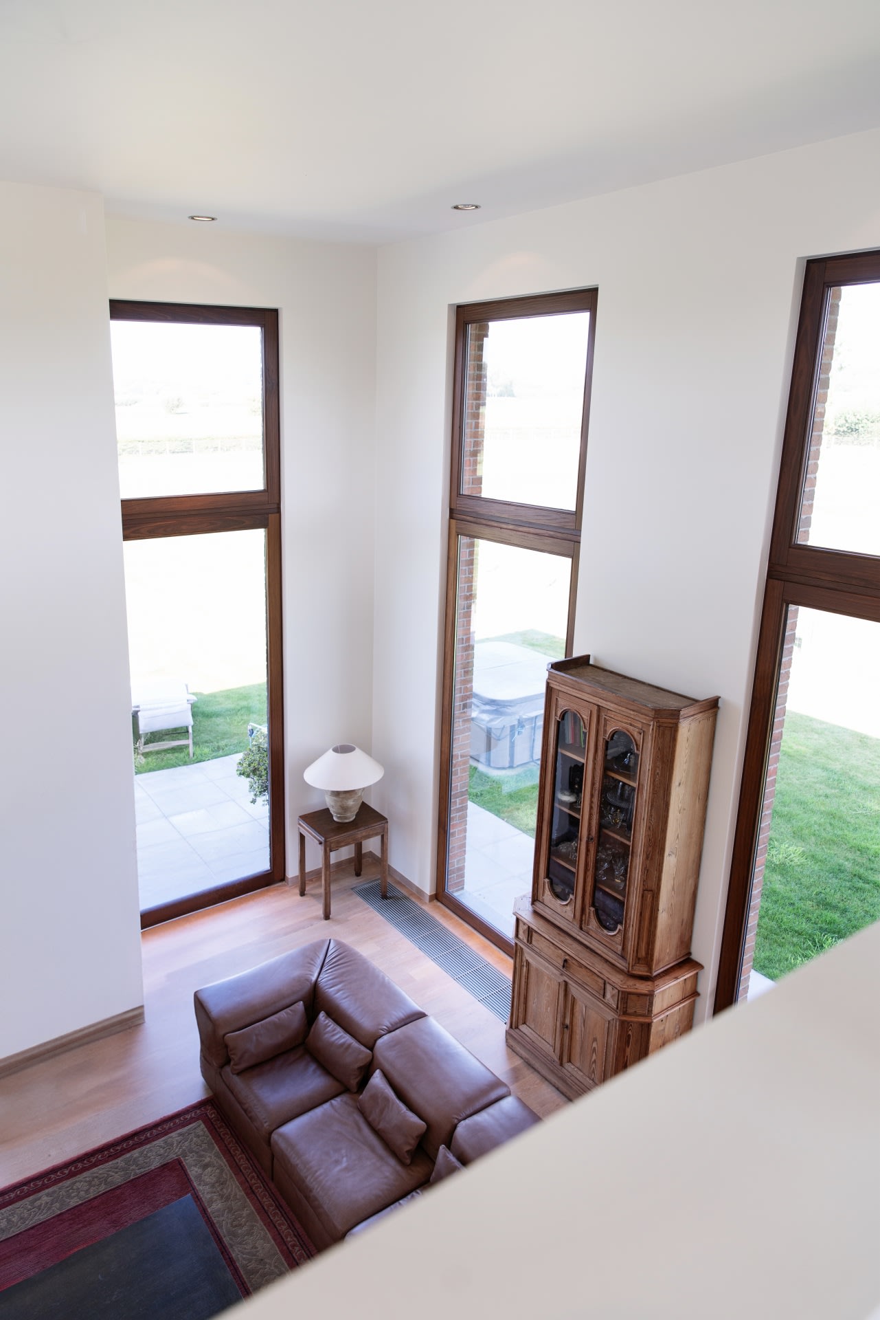 Un salon avec de hautes fenêtres, un canapé en cuir marron, une armoire en bois, une table d'appoint avec une lampe blanche et un tapis à bordures rouges, vu d'en haut.