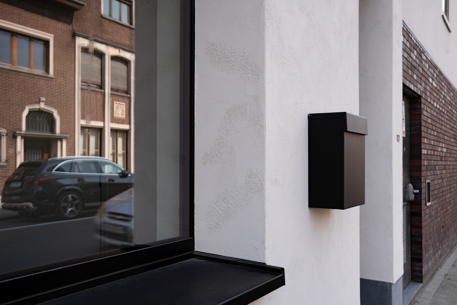 Een zwarte brievenbus is gemonteerd op een witte muur naast een groot raam, waarin een geparkeerde auto en gebouwen aan de overkant van de straat worden weerspiegeld.