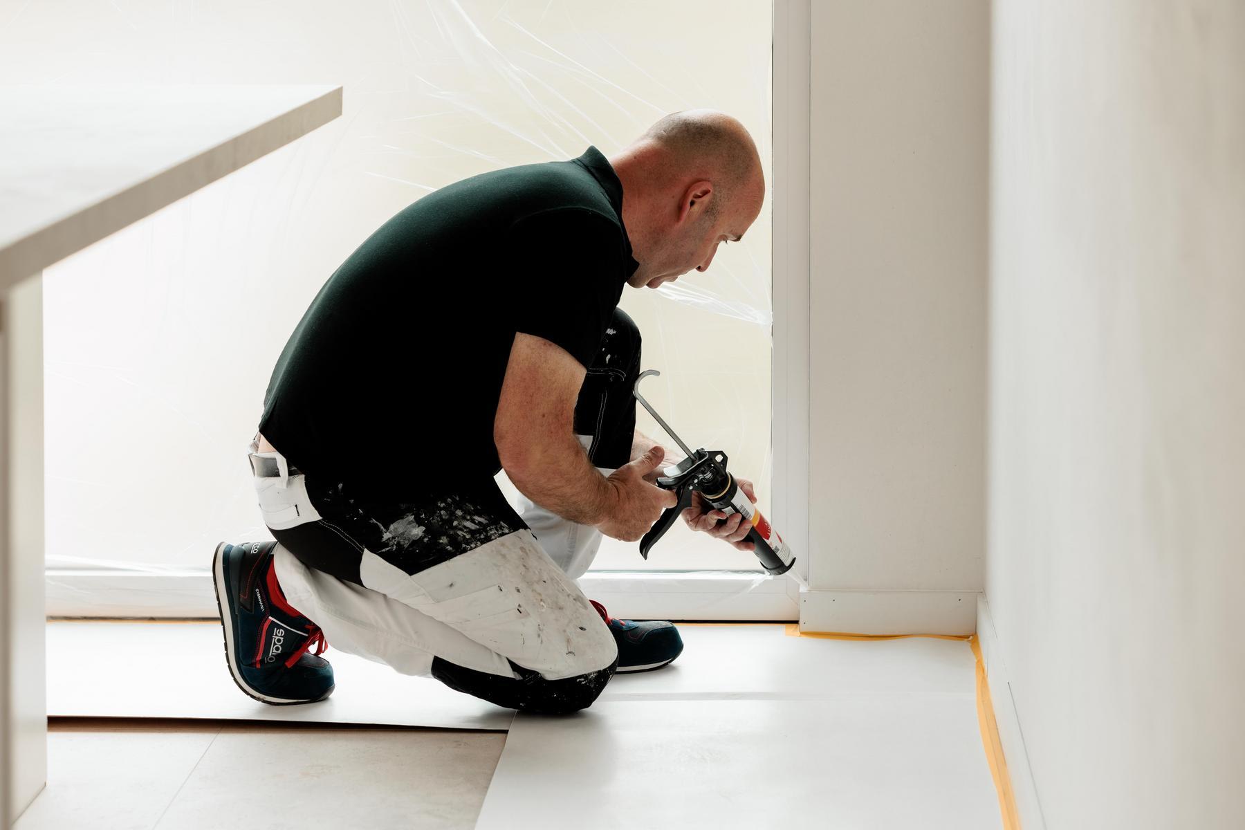 Een man knielt op de vloer en gebruikt een kitpistool om de randen tussen de vloer en de muur af te dichten in een moderne, lichte kamer.