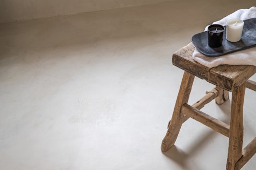 Een rustiek houten krukje houdt een zwart dienblad met een zwarte mok, een witte kaars en een witte doek vast, tegen een effen lichtgekleurde achtergrond van vloer en muur.