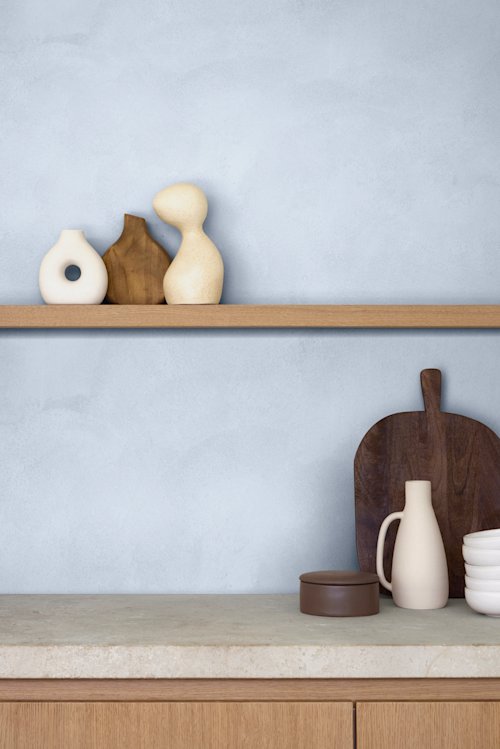 Een minimalistische keukenplank met drie decoratieve vazen, een houten snijplank, een crèmekleurige kan, gestapelde kommen en een ronde bruine bak tegen een lichtblauwe muur.