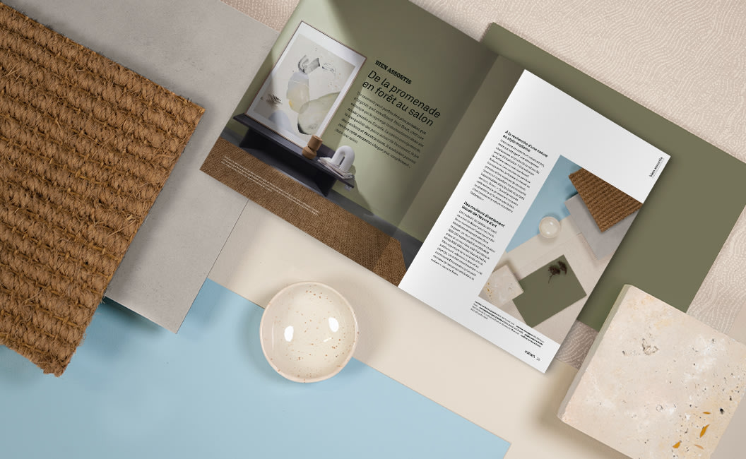Une composition à plat de matériaux d'intérieur, comprenant un échantillon de tissu texturé, un échantillon de pierre, un bol en céramique et un magazine colora ouvert sur un fond vert sauge et bleu.