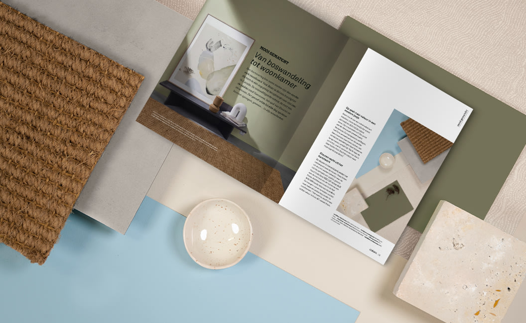 Een flat lay van interieurmaterialen waaronder een geweven textuurstaal, een steenstaal, een keramieken schaal en een open colora-magazine op een saliegroene en blauwe achtergrond.