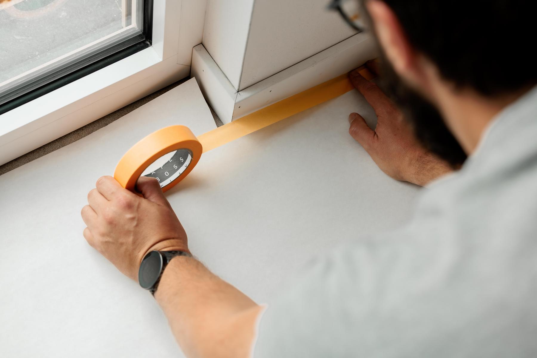 Een persoon plakt afplaktape langs de rand van een raam en een muur om het oppervlak voor te bereiden op het schilderen.