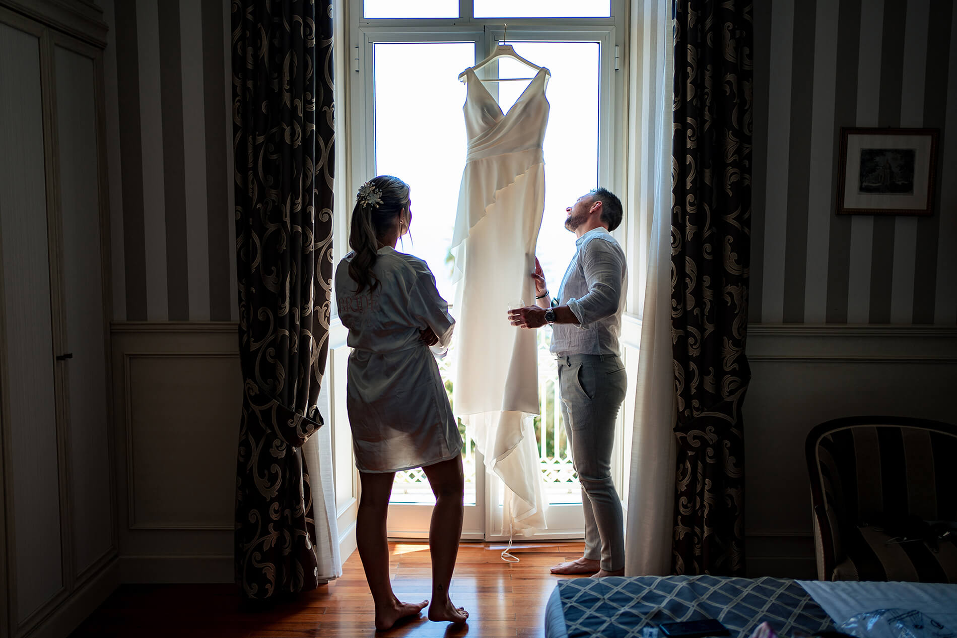 la sposa ed il testimone guardano il vestito da sposa appeso alla finestra all'hotel royal di sanremo.