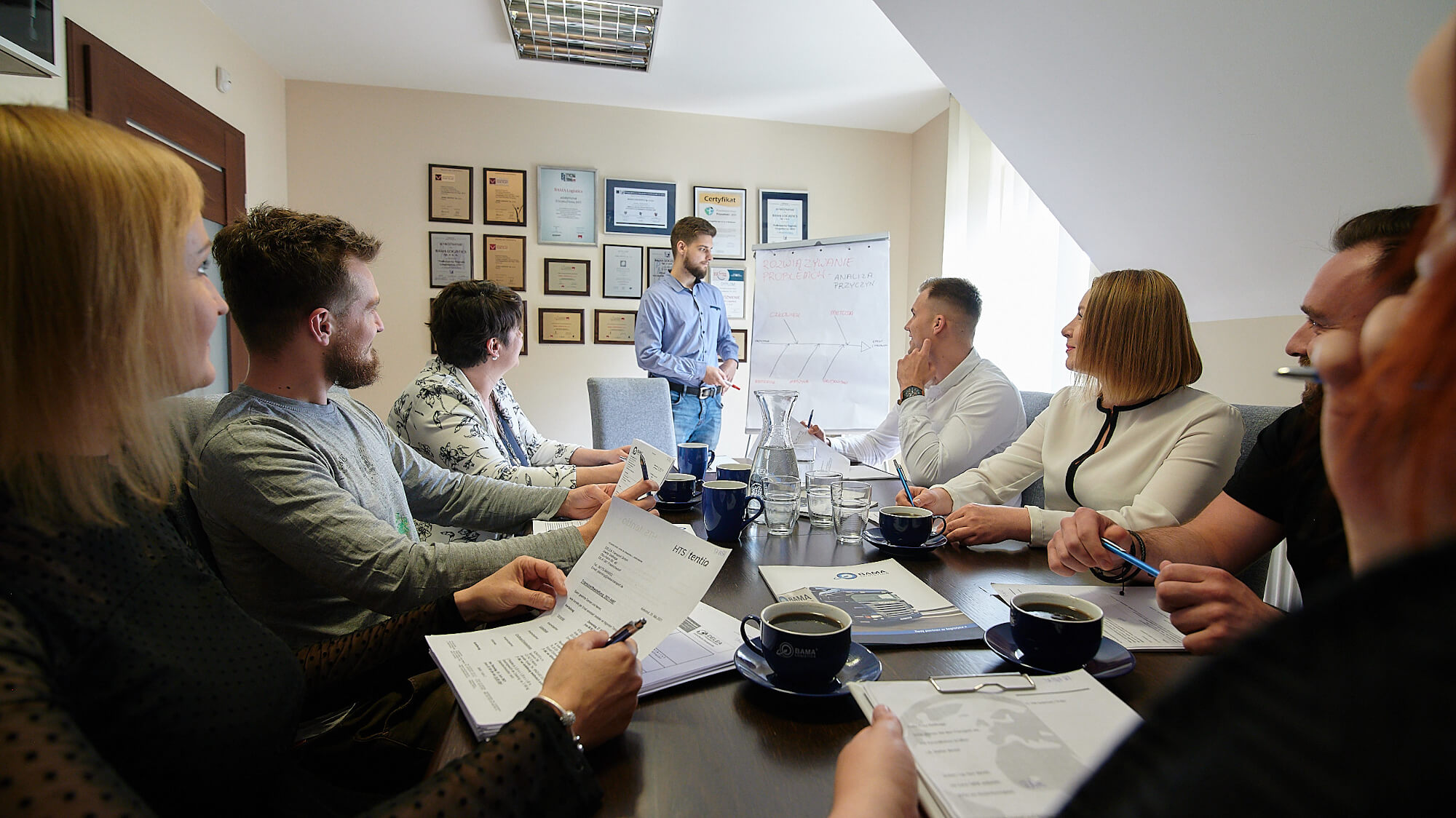 Narada przedstawicieli poszczególnych zespołów Firmy Bama Logistics w jej siedzibie w Kozłowie.  W tle certyfikaty potwierdzające kompetencje firmy i jej zespołu.