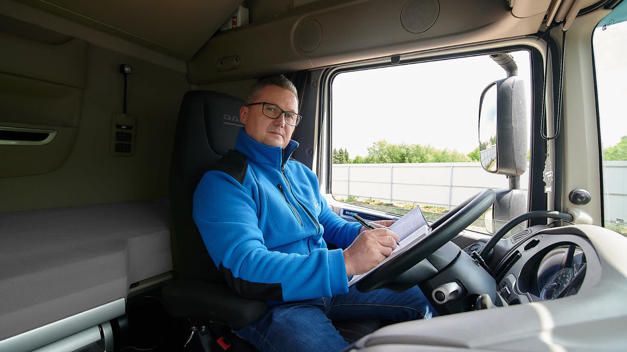 Kierowca, w stroju roboczym firmy Bama Logistics, siedzący w kabinie samochodu ciężarowego.