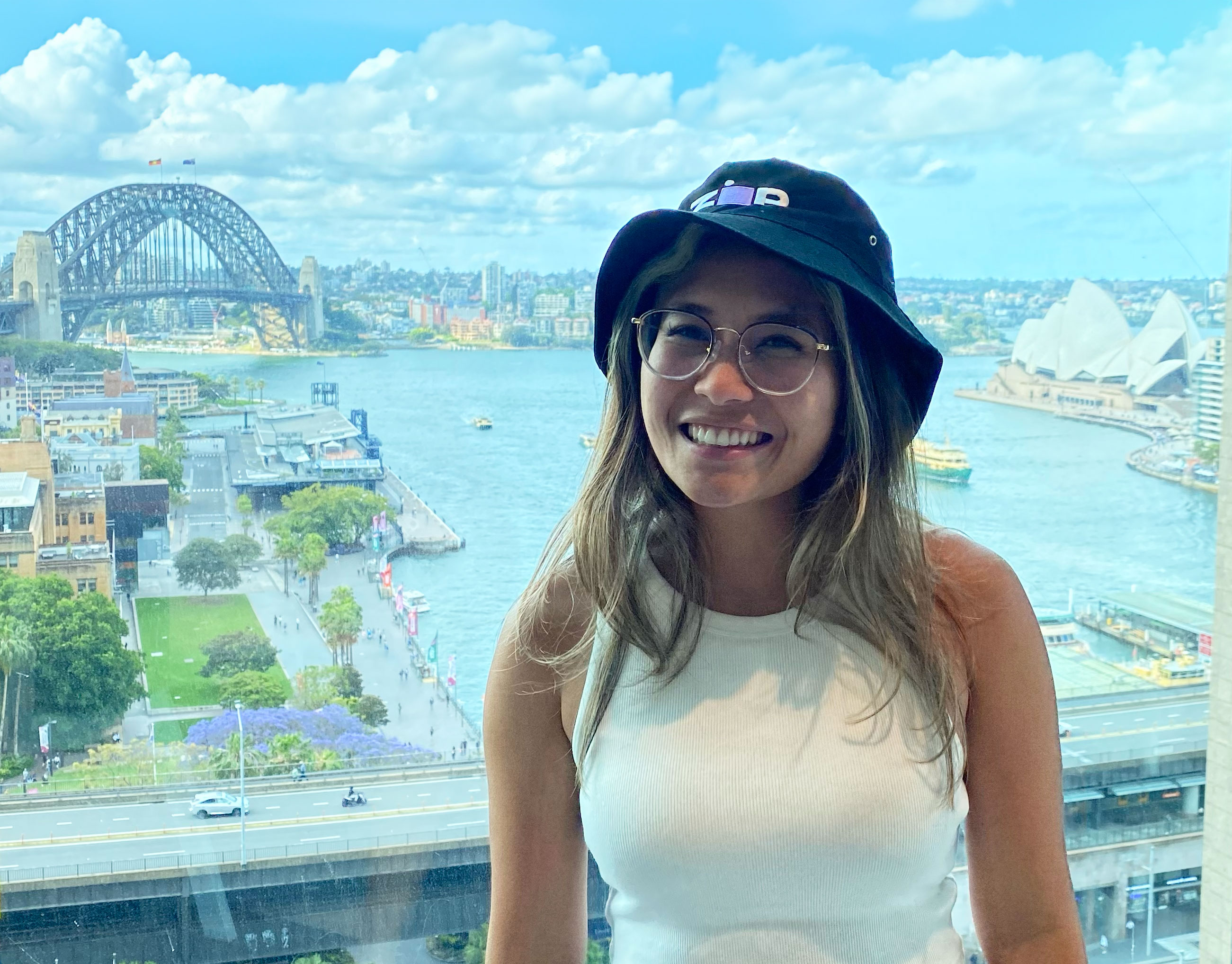 Sieny is wearing a white top and a #000000 Zip bucket hat with a backdrop of Sydney Harbour Bridge and the Opera House