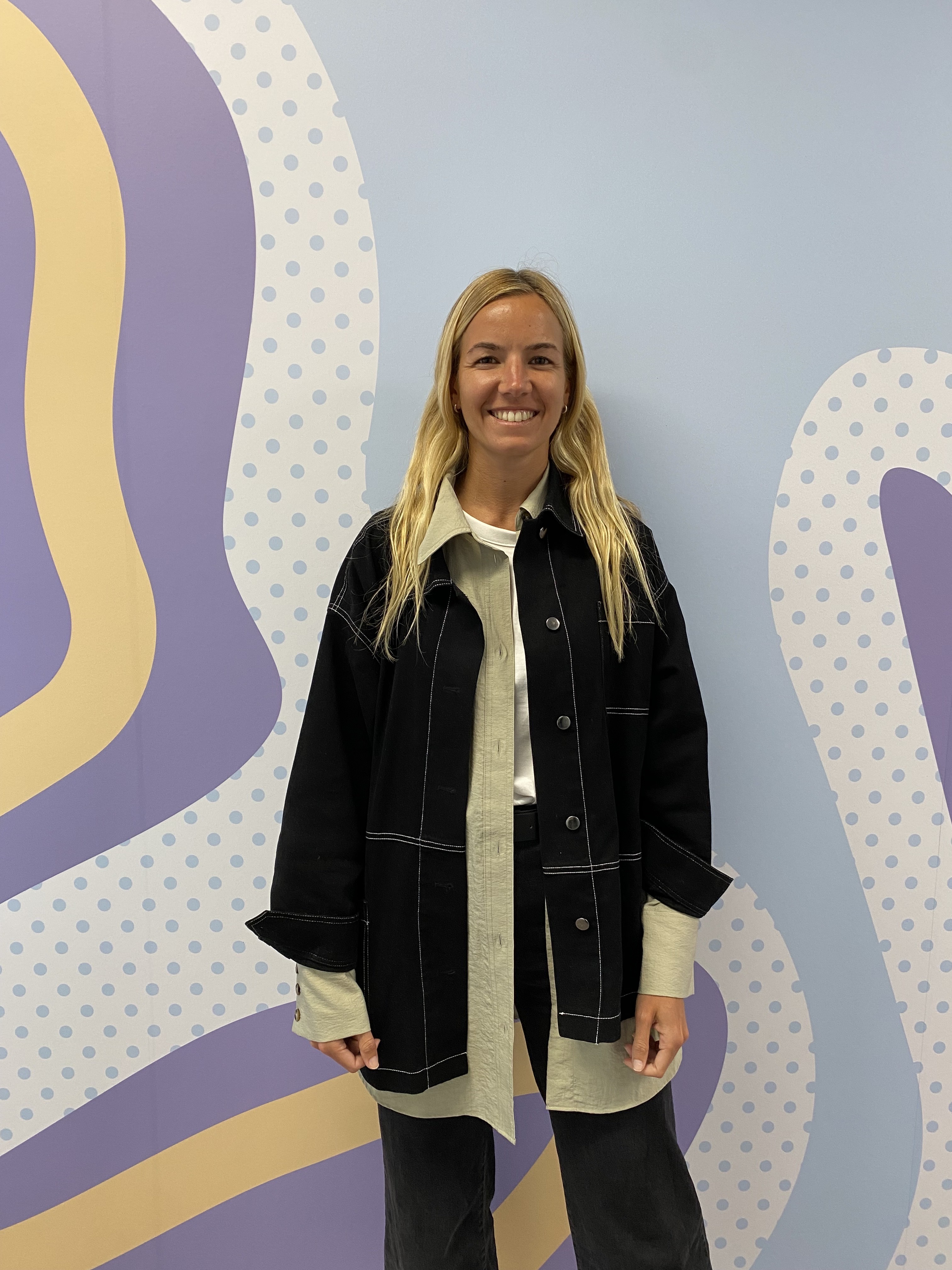 Clara is wearing a navy jacket and stands in front of a purple and blue Zip branded mural wall