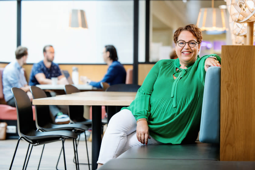 CHRO Suzanne van Niekerk zit op een bankje op kantoor en lacht naar de camera. Achter haar is een drietal collega's vervaagd zichtbaar. 