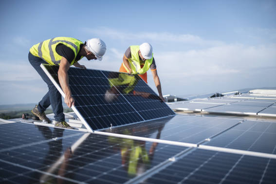 Two workers wearing high-visibility vests and hard hats, standing on a roof and installing solar panels.