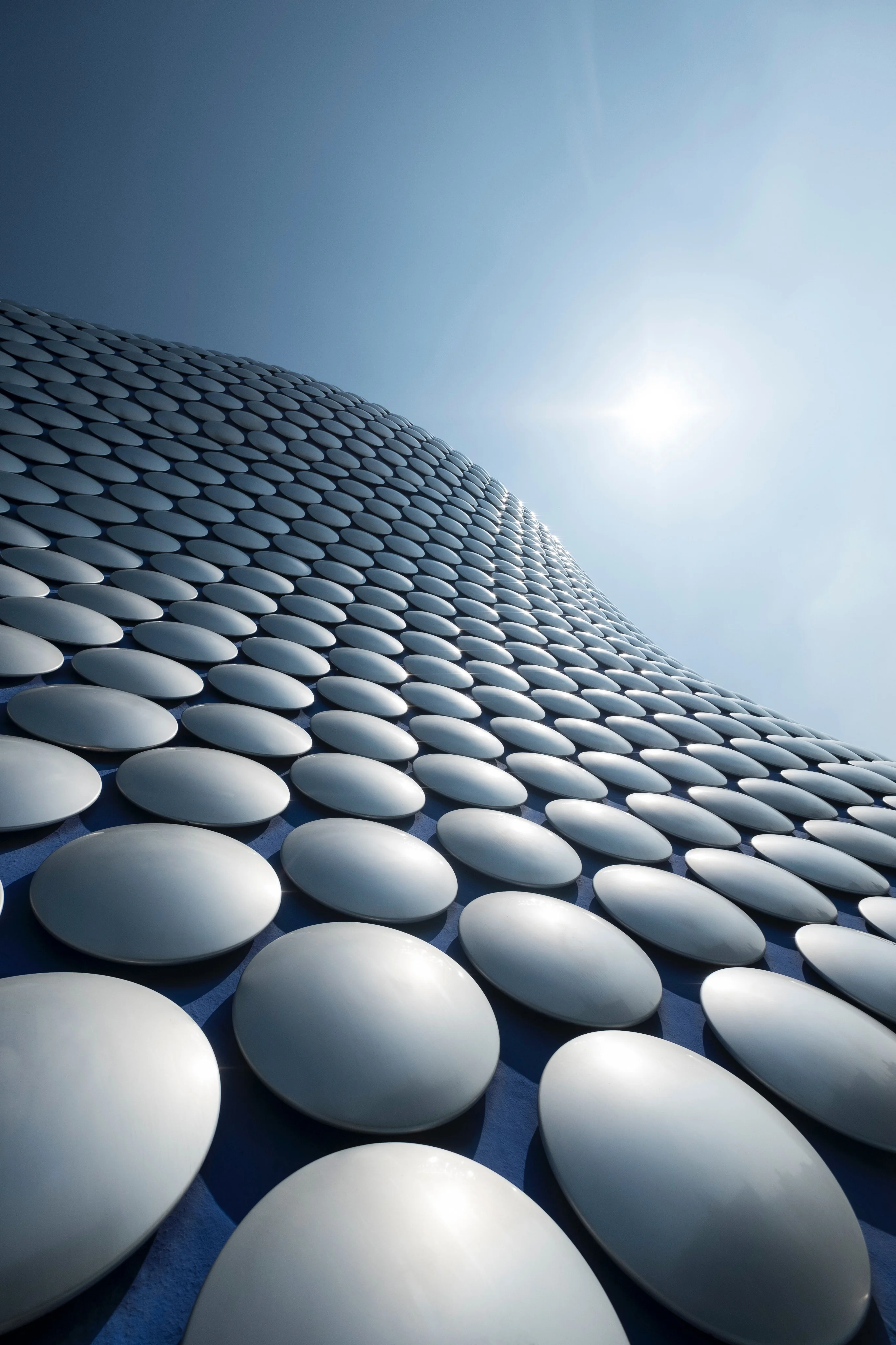 An image of the outside of selfridges in birmingham from below looking up. You can see the iconic silver circles with the blue background of the building and the sky on a sunny day