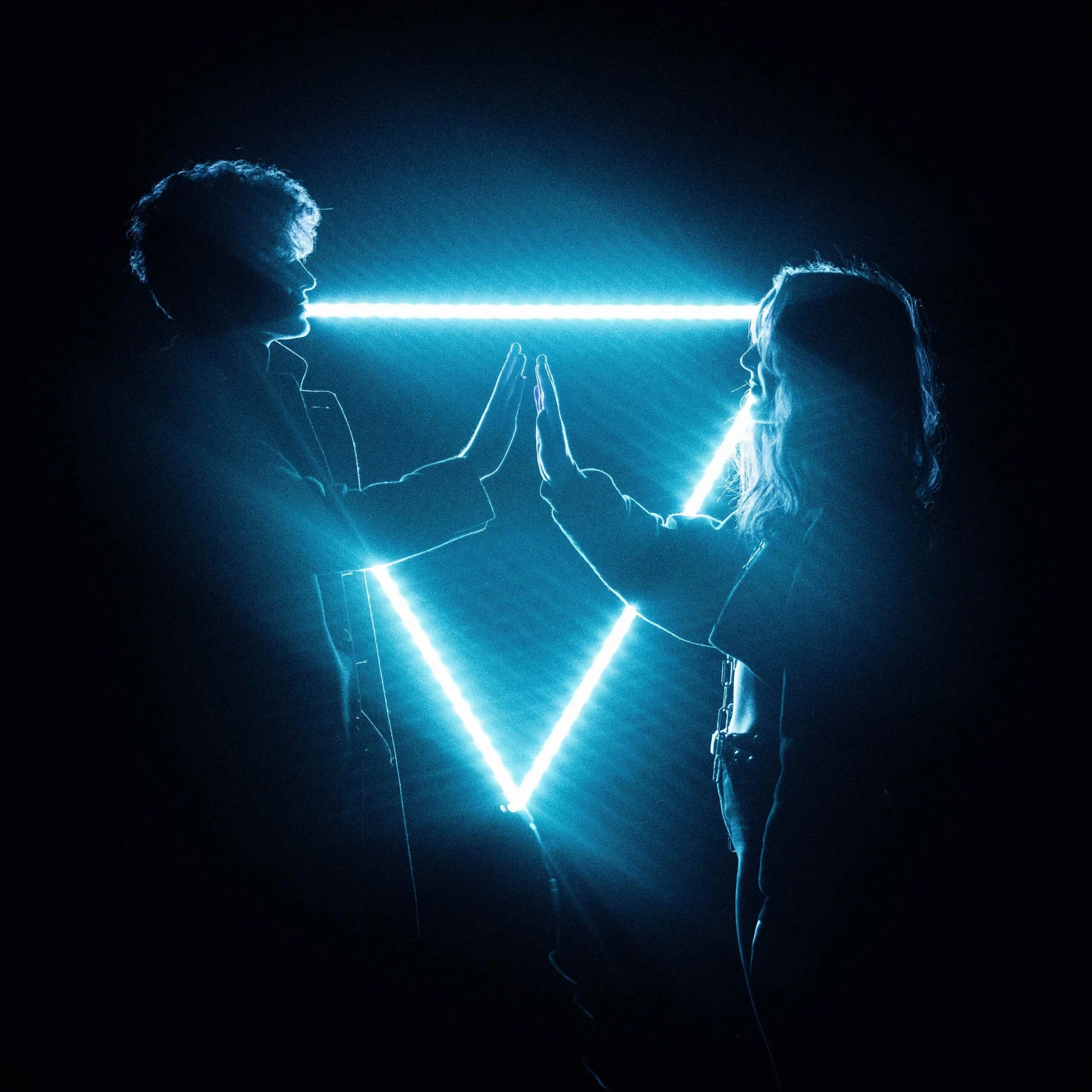 2 people facing each other with one of their hands up, about to touch the other persons hand. Upside-down triangle light in the background