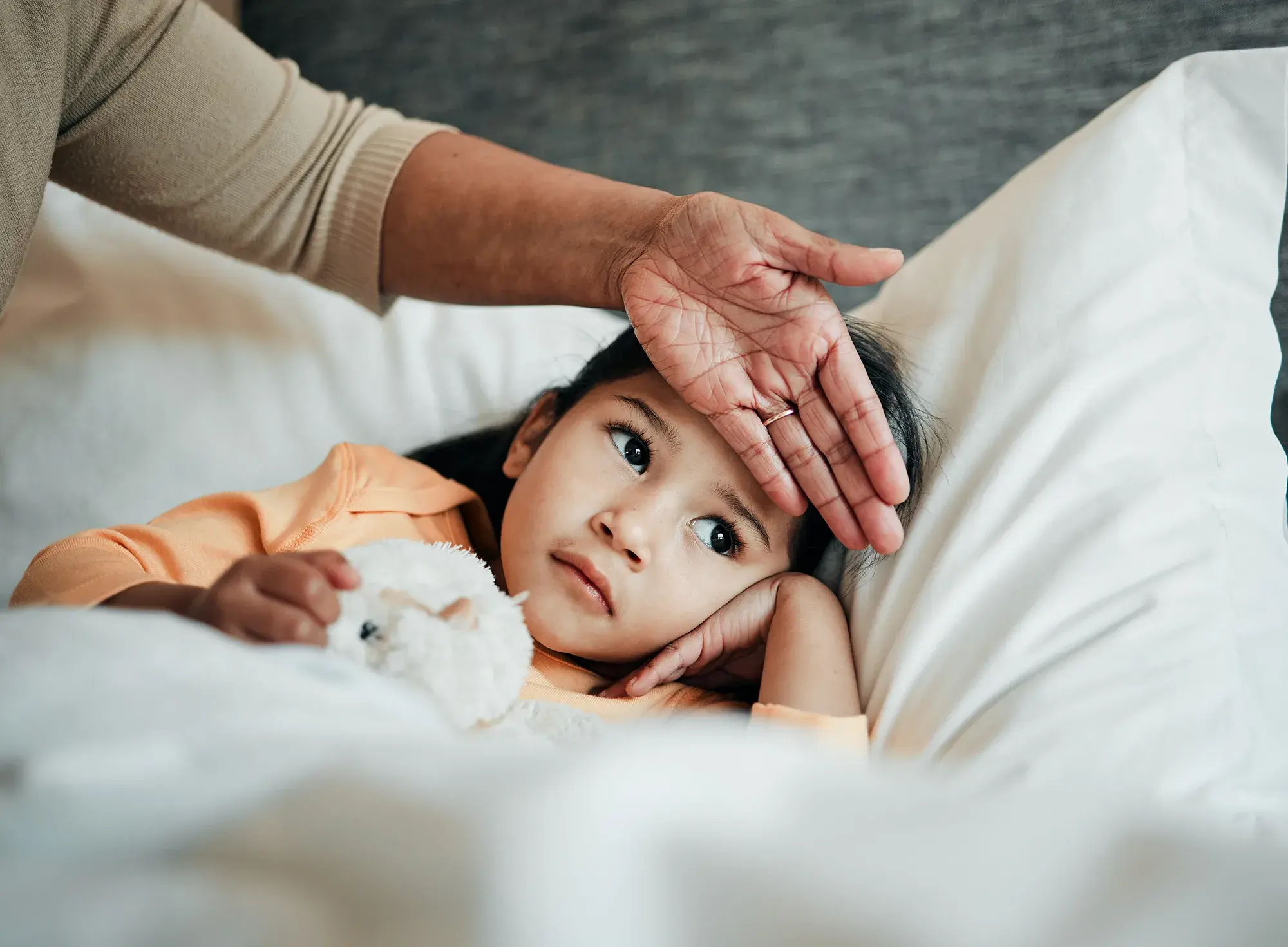 A young child lying in bed with a parent’s hand gently resting on their forehead to check for a fever