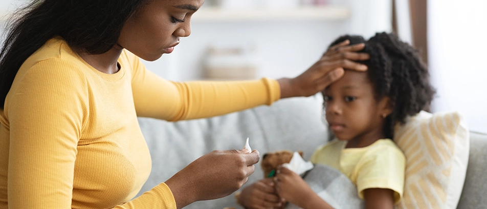 A mother is sitting on a couch with her young daughter, placing one hand on the child’s forehead, while her other hand holds a thermometer. The mother looks at the thermometer with concern, while the daughter is holding a teddy bear and a tissue.