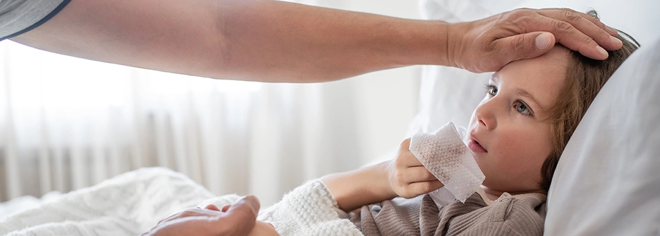 A young girl is lying in bed with the covers over her and a tissue in her hand. An adult man’s arm is seen in the photo with his hand on the child’s head feeling for a temperature. His other hand is holding the little girl’s other hand.