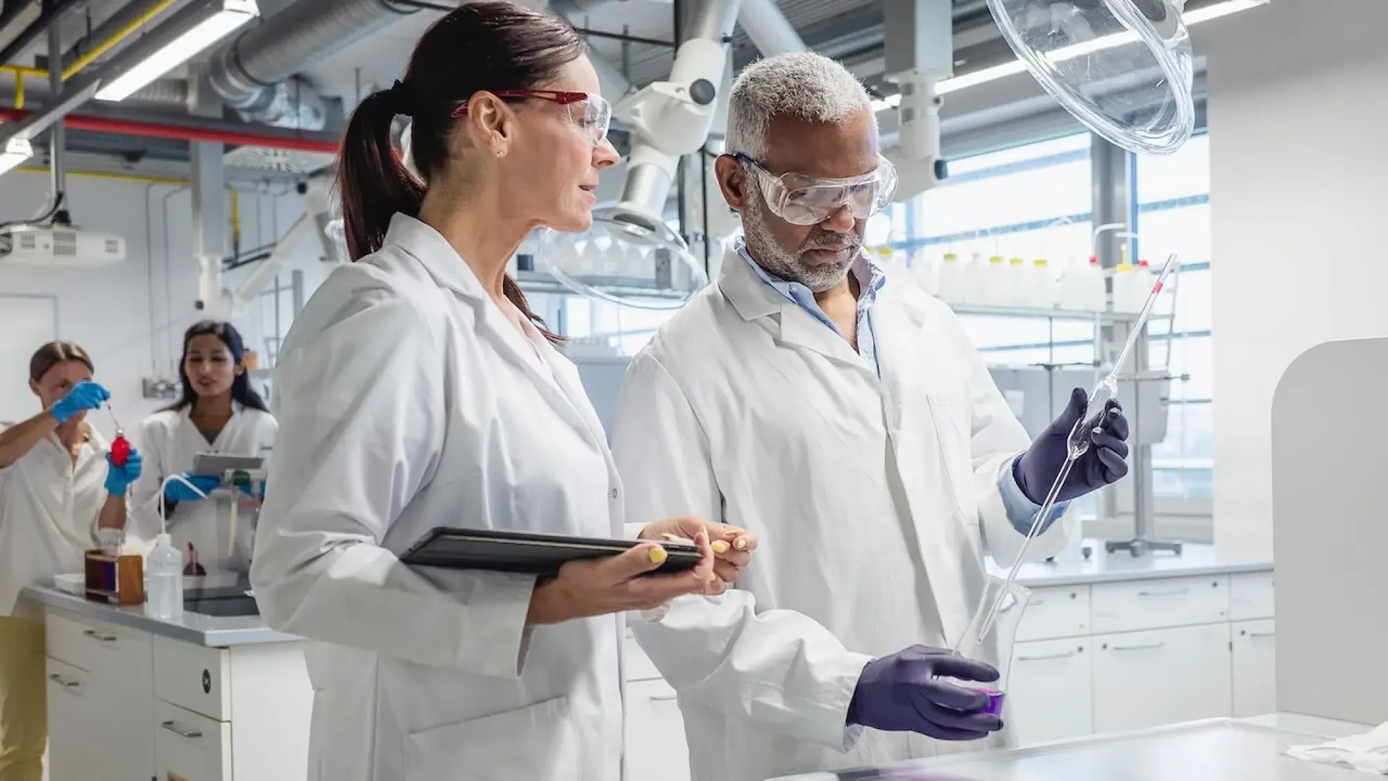 A diverse team of four laboratory scientists wearing lab coats and safety goggles works together in a modern research lab, with two researchers in the foreground examining lab equipment and two scientists using lab equipment in the background.