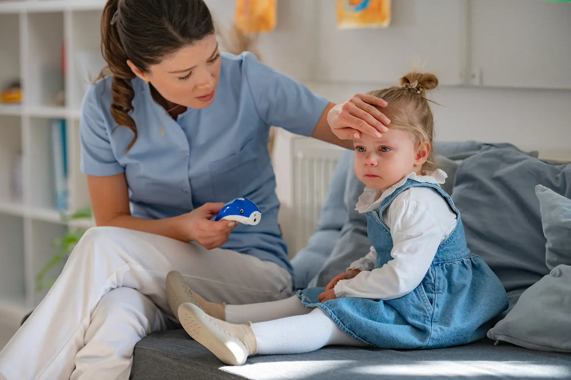 A mother sits with her sick toddler on a couch, holding a thermometer in one hand and placing her other hand on the young girl's forehead. The child is sitting up and looks sad, while the mother appears concerned.