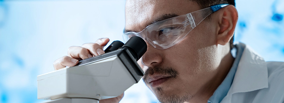 A male laboratory scientist wearing safety glasses and a lab coat looks through a microscope while holding the top of it with one hand. A large screen can be seen in the background.