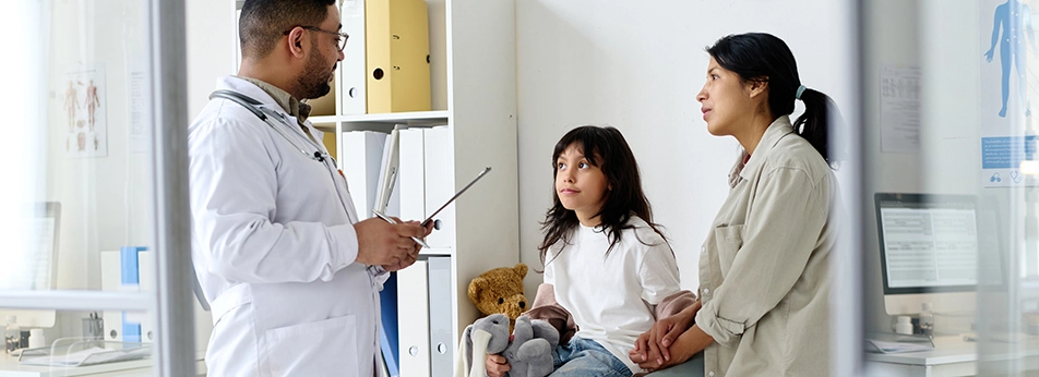 Mother and daughter discuss health with a doctor in an exam room; child holds a stuffed bunny.