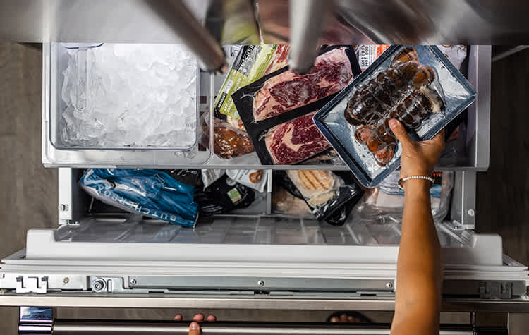 Hand reaching into freezer full of individually packaged meat, including lobster and steaks.