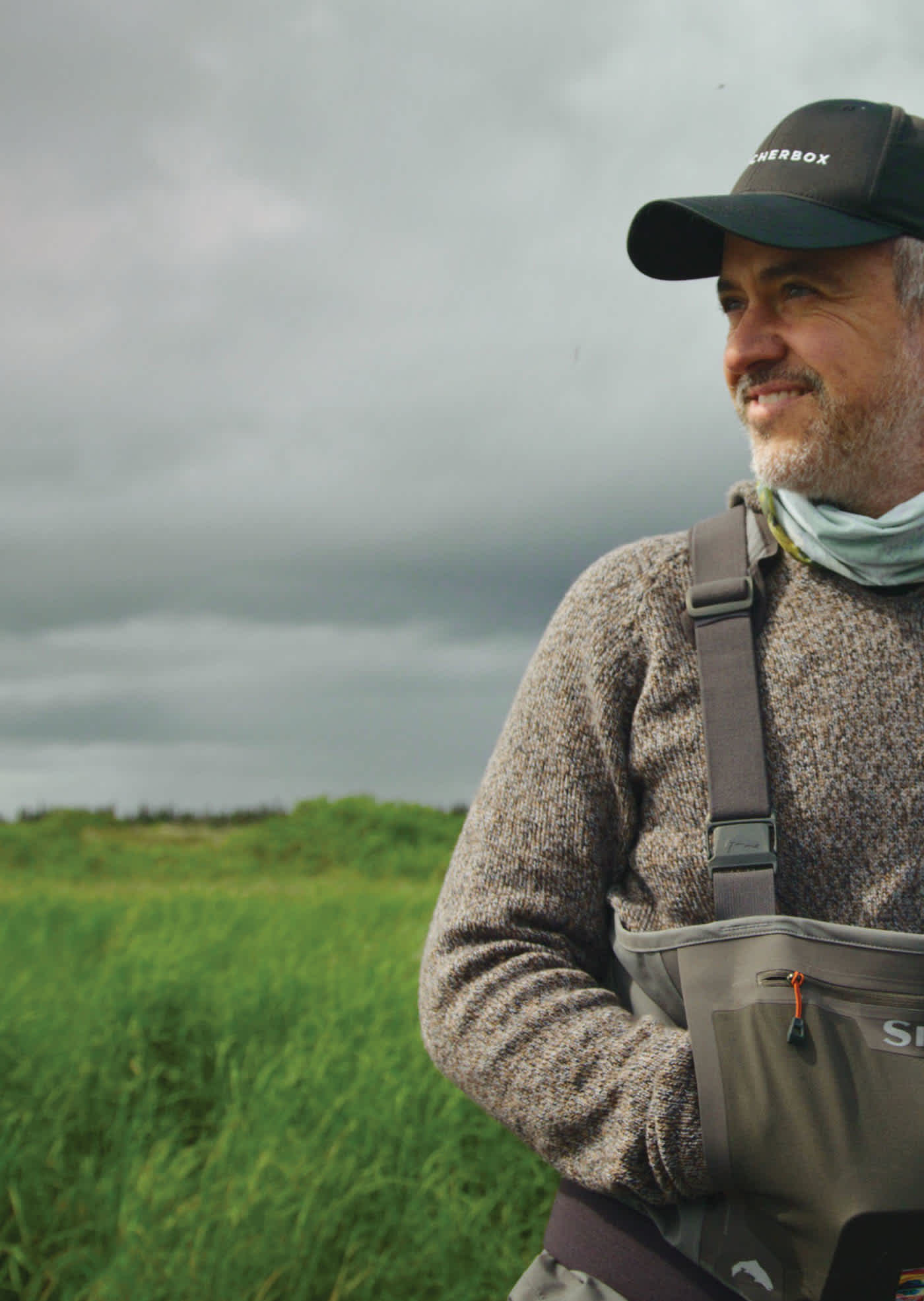 ButcherBox CEO, Mike, wearing a hat and sweater stands in a green field, surrounded by tall grass under a clear blue sky.