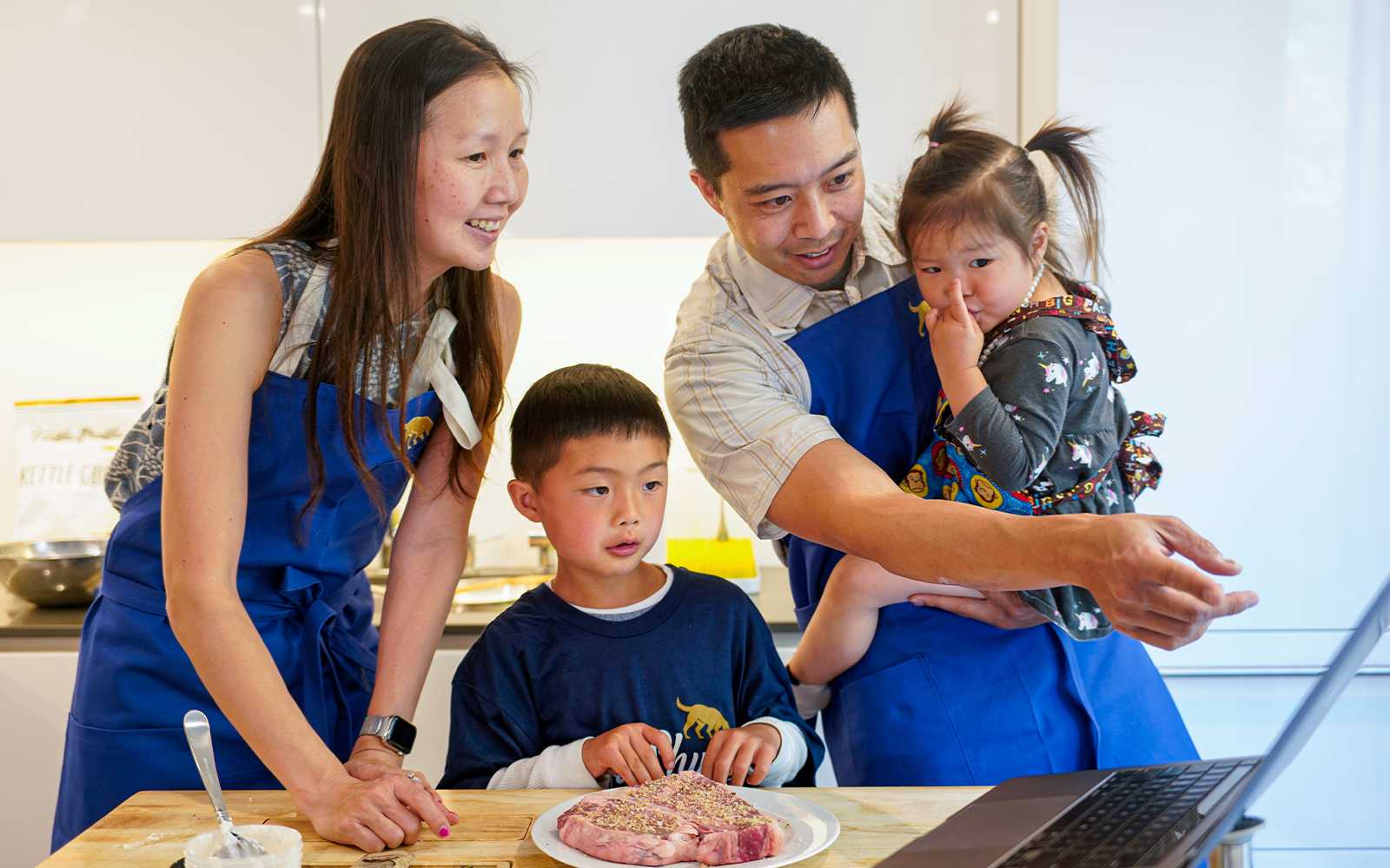 Family in the kitchen wearing truffle shuffle aprons, looking at a computer screen and making a meal together.