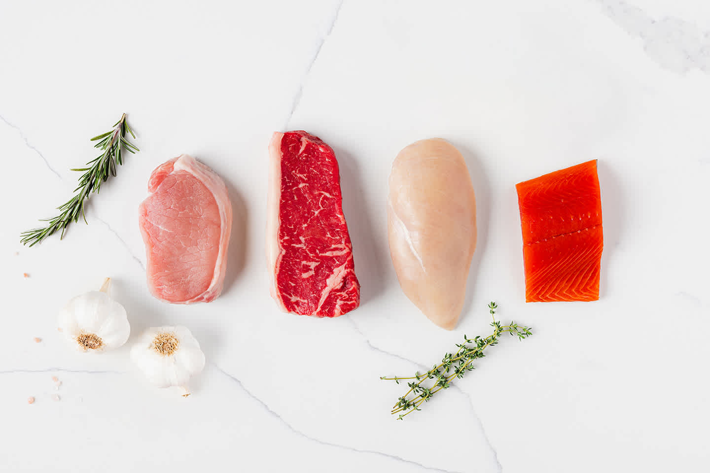 white marble background displaying four cuts of meat, pork, steak, chicken, and salmon, surrounded by herbs and garlic