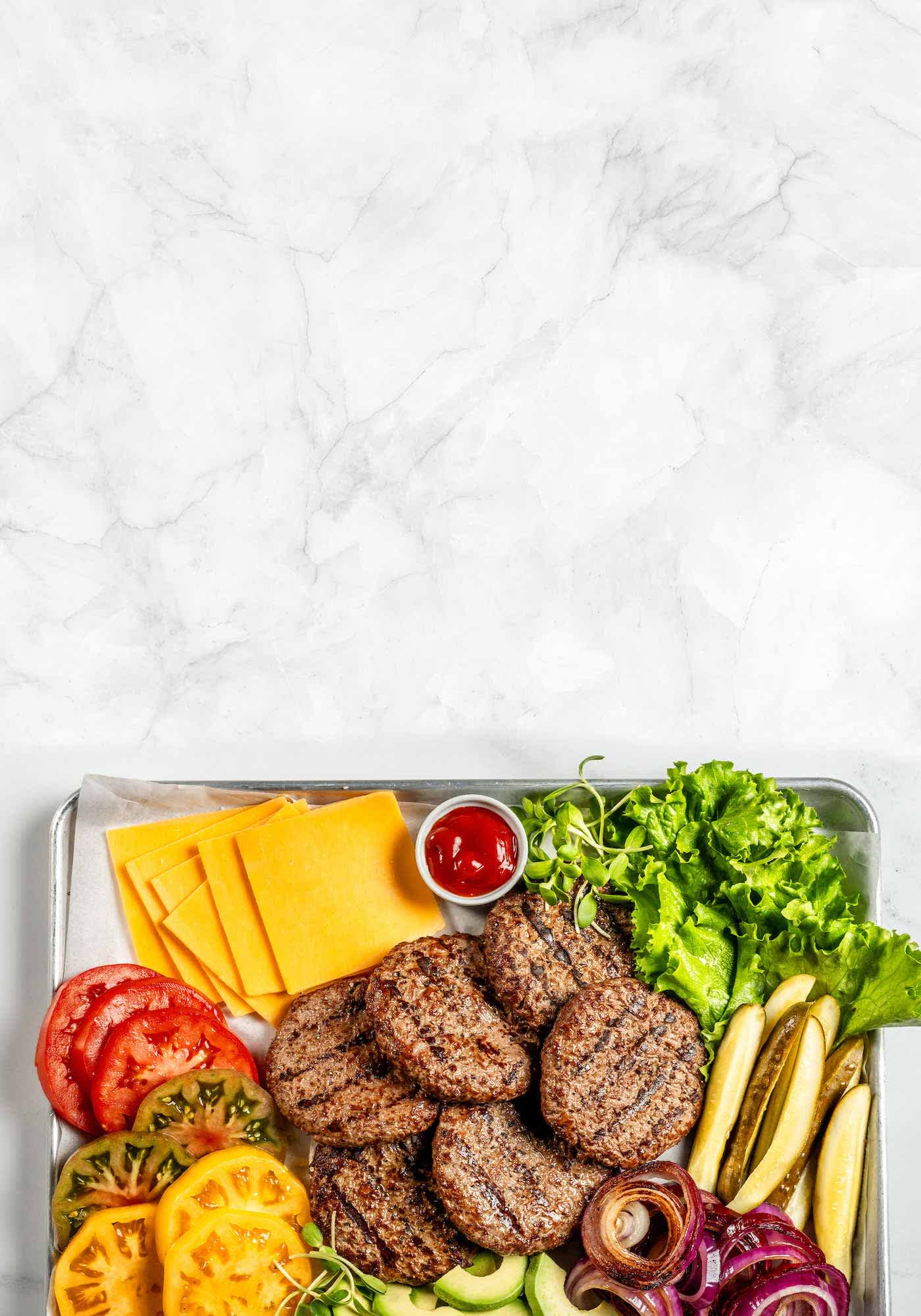 A tray of cooked burger patties and burger toppings on a marble background.