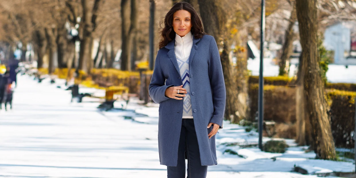 Ein Model mit dunklen Haaren trägt einen langen blauen Mantel, eine weiße Strickjacke und Jeans.