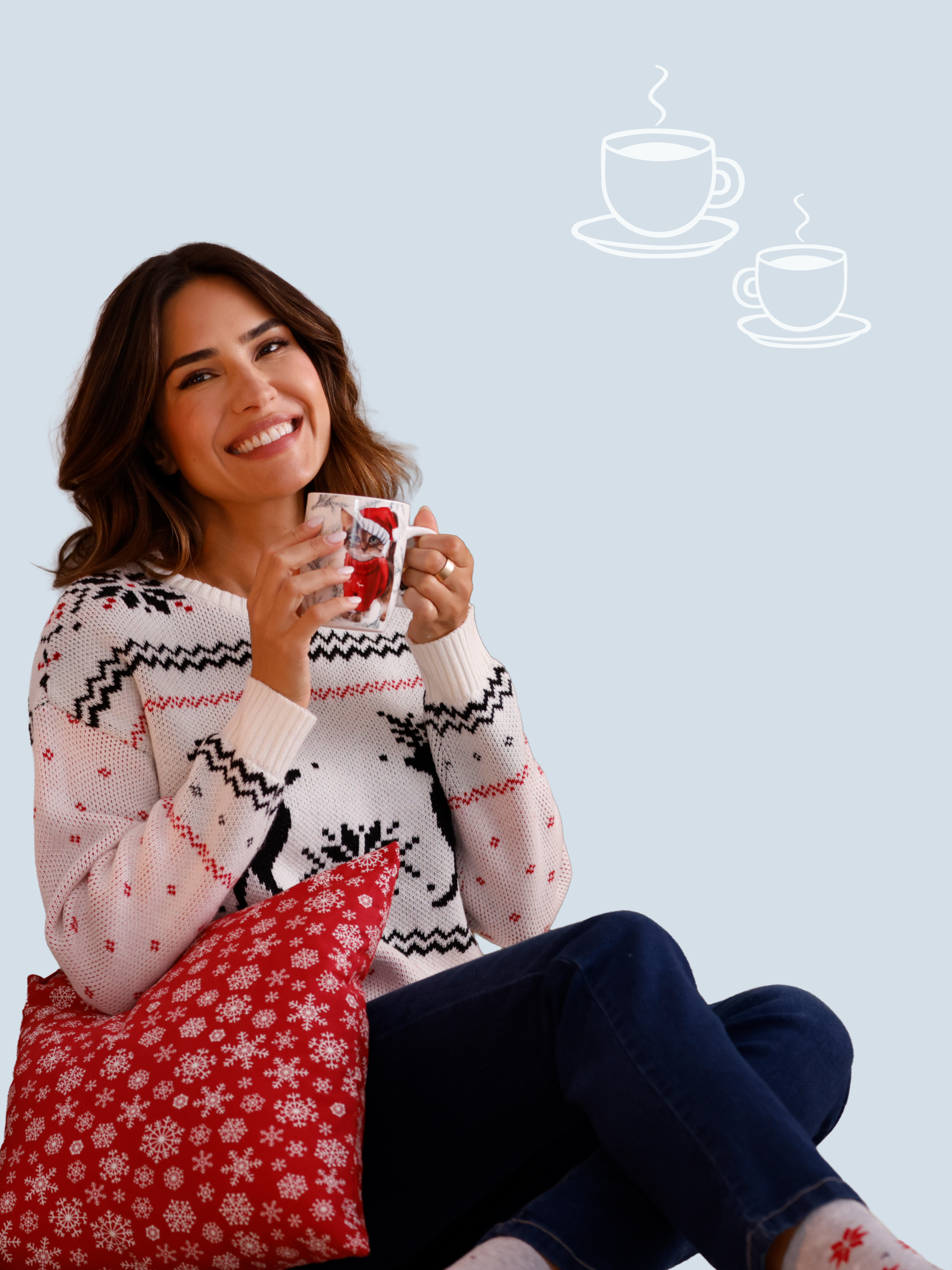 Een model draagt een witte trui met zwart rendierpatroon en houdt een mok vast. Zit met een rood kussen op schoot.