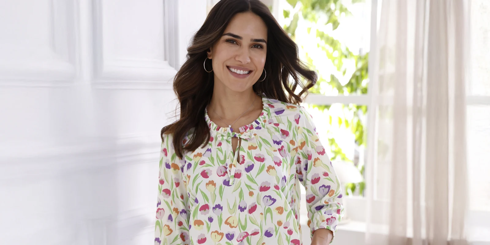 Modelka má na sebe bielu blúzku s farebným vzorom tulipánov a 3/4 rukávmi pred svetlým pozadím.