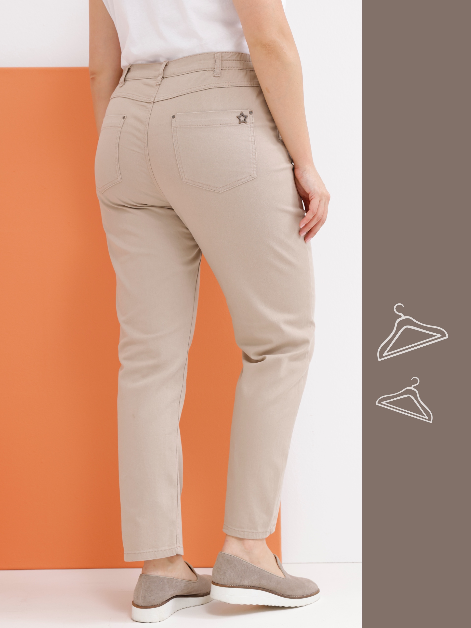 Ein Model von hinten in beigen Hosen und grauen Schuhen, vor einem orangefarbenen Hintergrund.