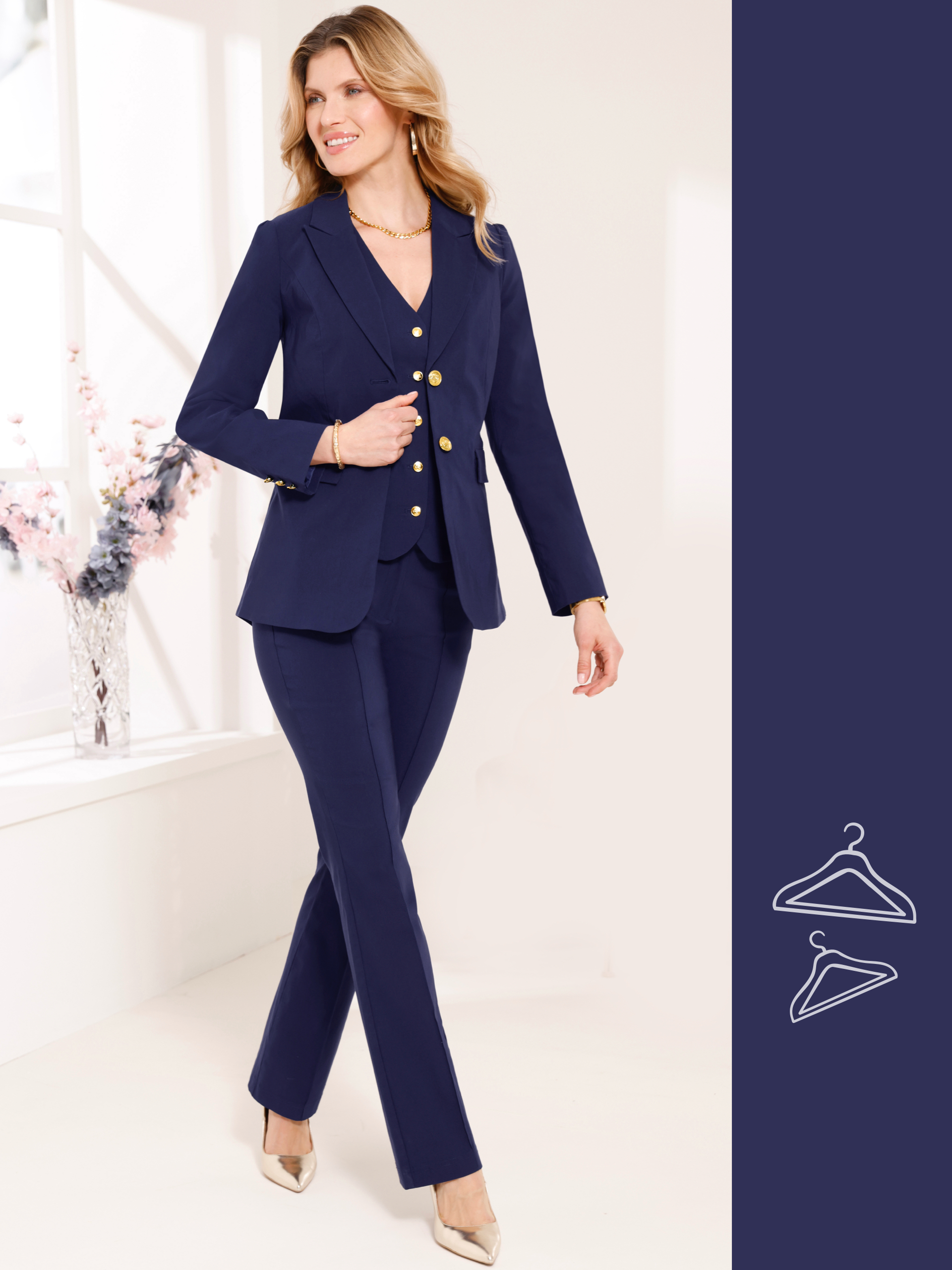 En modell med blont, vågigt hår bär en mörkblå kostym med guldknappar, ett guldfärgat halsband och guldfärgade skor.