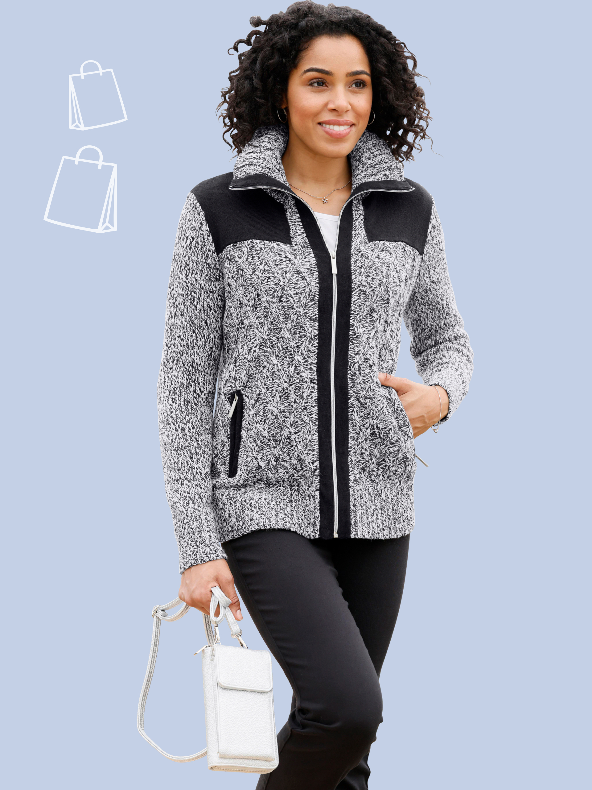 Ein Model trägt eine graue Strickjacke mit schwarzem Schulterbesatz und schwarzen Hosen, hält eine weiße kleine Tasche.