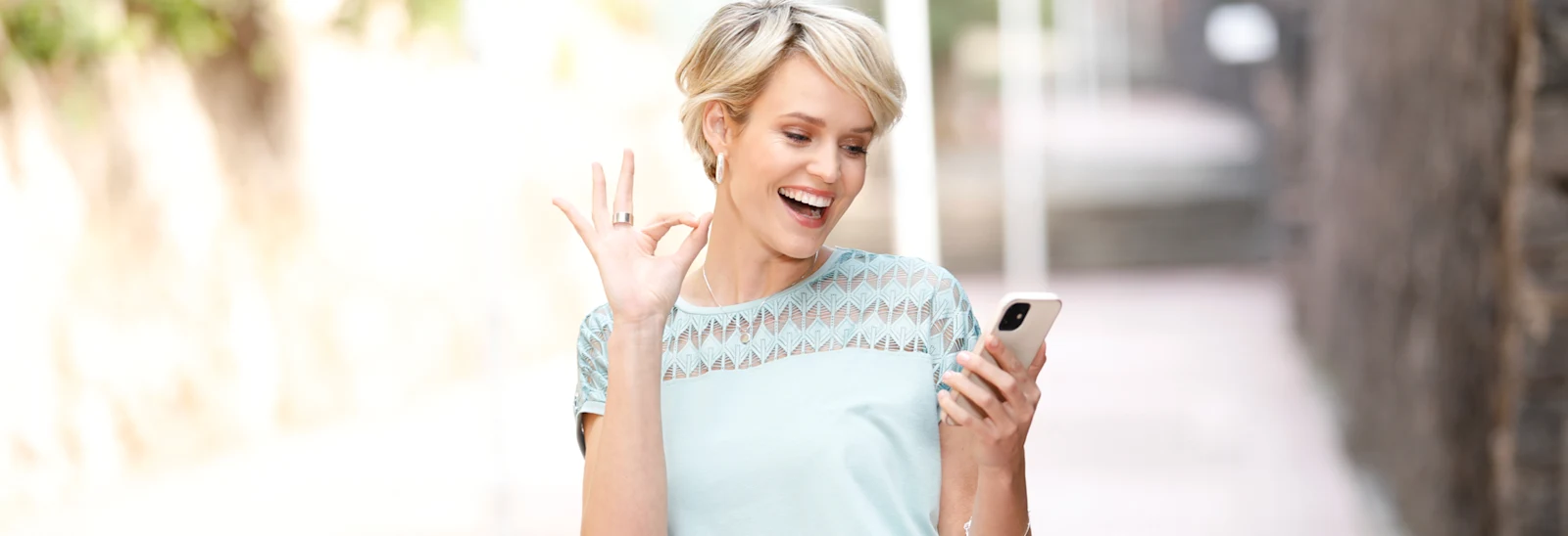 Kvinna i ljusblå topp håller en smartphone och gör en 'OK'-gest med handen, utomhus i en solbelyst miljö. Kvinna i ljusblå topp håller en smartphone och gör en 'OK'-gest med handen, utomhus i en solbelyst miljö.