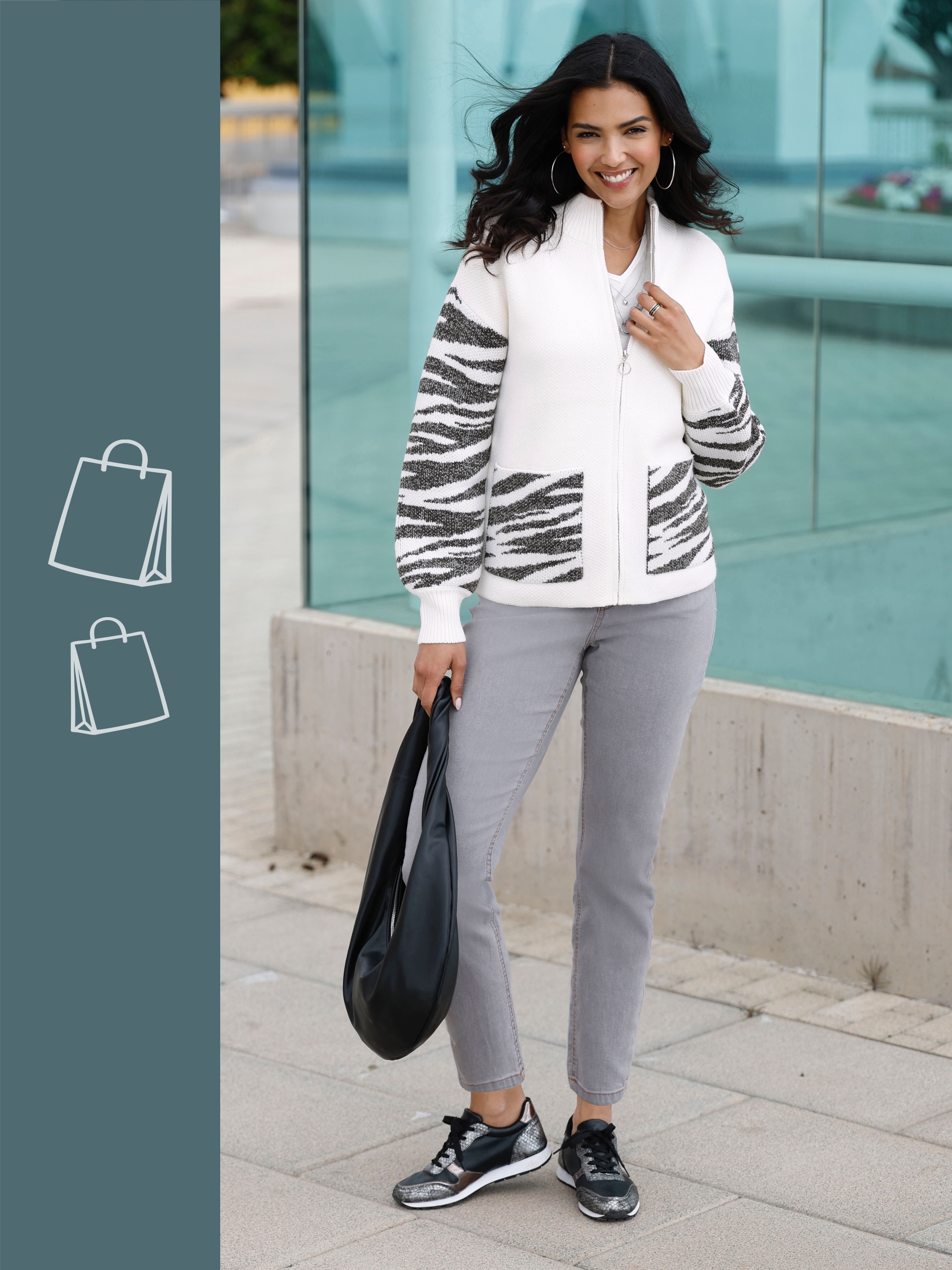 Ein Model trägt einen weißen Cardigan mit gestreiften Ärmeln, graue Hosen und schwarze Sneakers. Es hält eine schwarze Tasche.