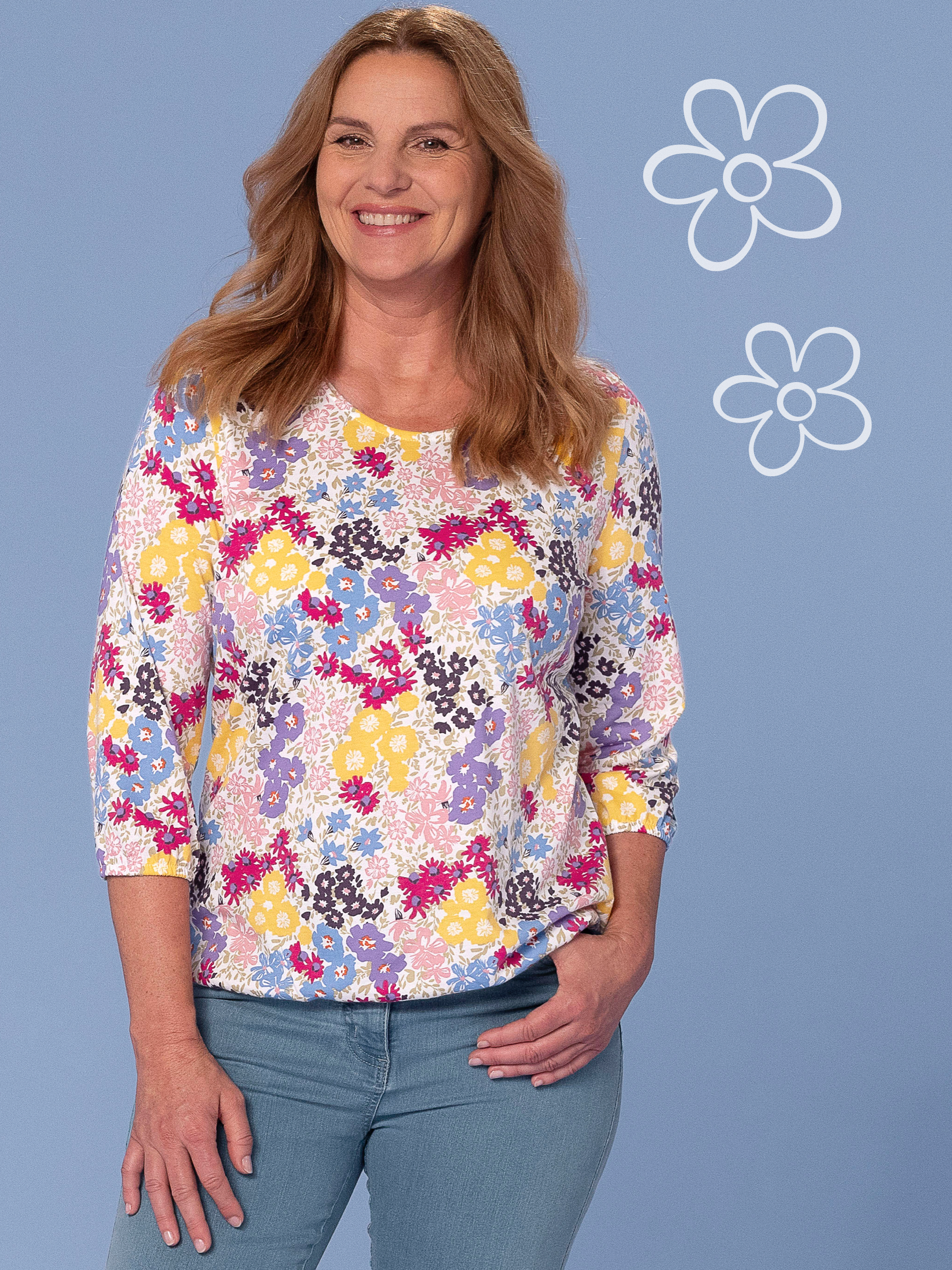 Damen-Shirt mit buntem Blumenmuster, 3/4-Ärmeln und Rundhalsausschnitt, kombiniert mit hellblauer Jeans vor blauem Hintergrund.