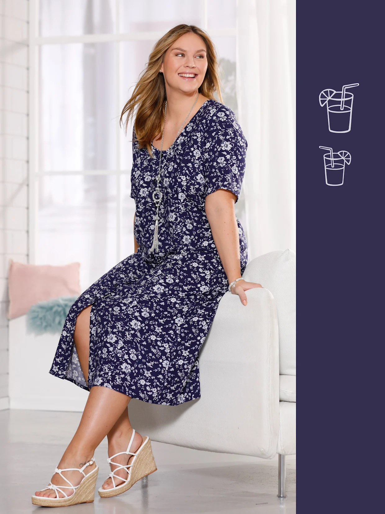 Ein Model sitzt auf einem weißen Sofa und trägt ein blumiges, dunkelblaues Kleid mit Wedgeschuhen.